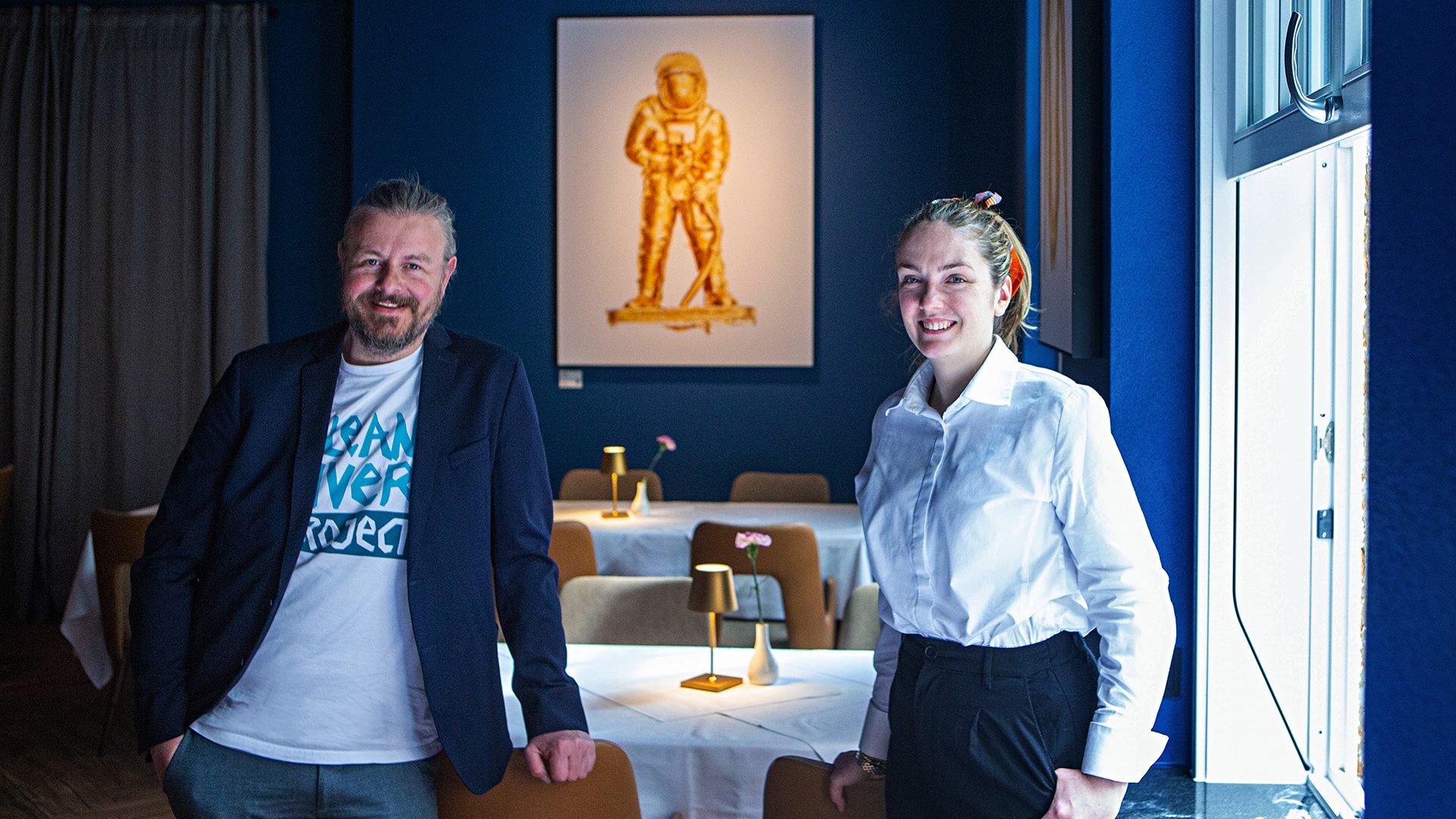 Stephan Horch und Naomi Burgardt im BEES Restaurant. Das Kunstwerk "Astronaut" leuchtet gold im Hintergrund.